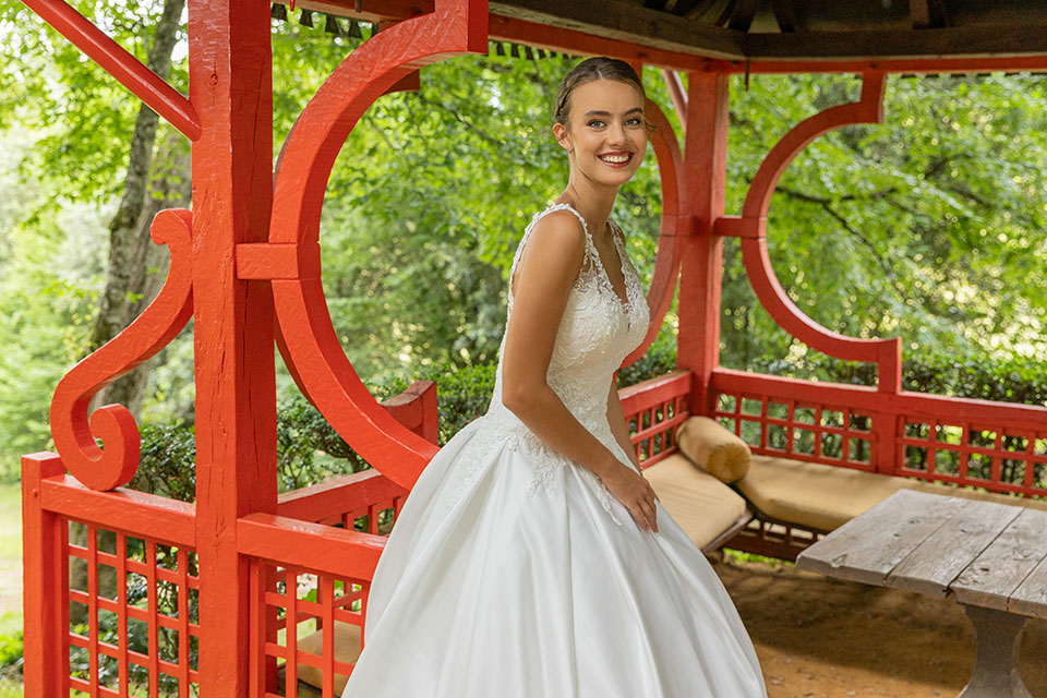 Bruid in een witte jurk met kanten details lacht terwijl ze in een sierlijke rode pergola staat omgeven door groene bomen.