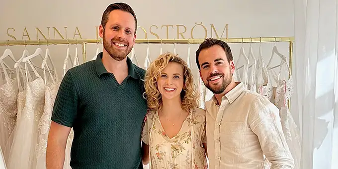 Twee mannen en een vrouw staan glimlachend voor een rek met trouwjurken. De man links draagt een donkergroene polo en kaki broek, de vrouw in het midden een bloemenjurk, en de man rechts een beige linnen overhemd en bijpassende broek. Het interieur is stijlvol en licht, met de naam 'SANNA LINDSTRÖM' in gouden letters op de muur, wat de sfeer van een bruidswinkel benadrukt.