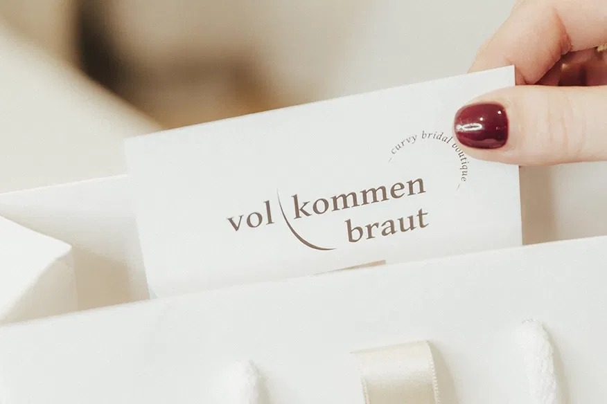 Eine Hand hält eine Visitenkarte mit der Aufschrift "vollkommen braut - curvy bridal couture" vor einem unscharfen, hellen Hintergrund.