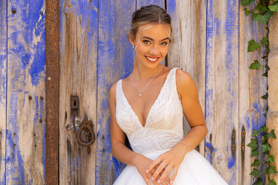 Gelukkige bruid met een brede glimlach in een witte trouwjurk staat voor een verweerde blauwe deur met ijzerbeslag.