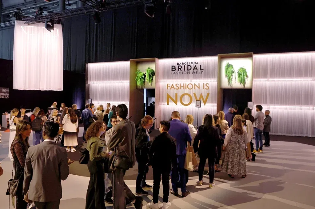 De ingang van de Barcelona Bridal Fashion Week met een moderne, verlichte entree en een banner met "FASHION IS NOW", waar een menigte van bezoekers samenkomt.