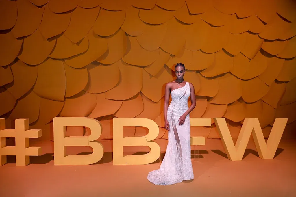 Elegante bruid poseert met een asymmetrische avondjurk tijdens Barcelona Bridal Fashion Week, gehuldigd door een oranje geometrische achtergrond met de hashtag #BBFW.