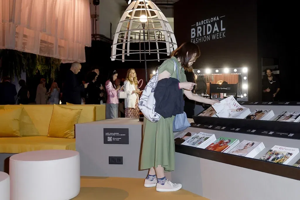 Bezoekers bekijken trouwmagazines bij een stand tijdens de Barcelona Bridal Fashion Week, verlicht door sfeervolle gele en witte lampen.