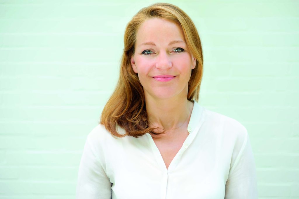 Porträtfoto von Barbara Koch, lächelnde Frau mit mittellangem, welligem Haar, trägt eine weiße Bluse, mit hellem Hintergrund.
