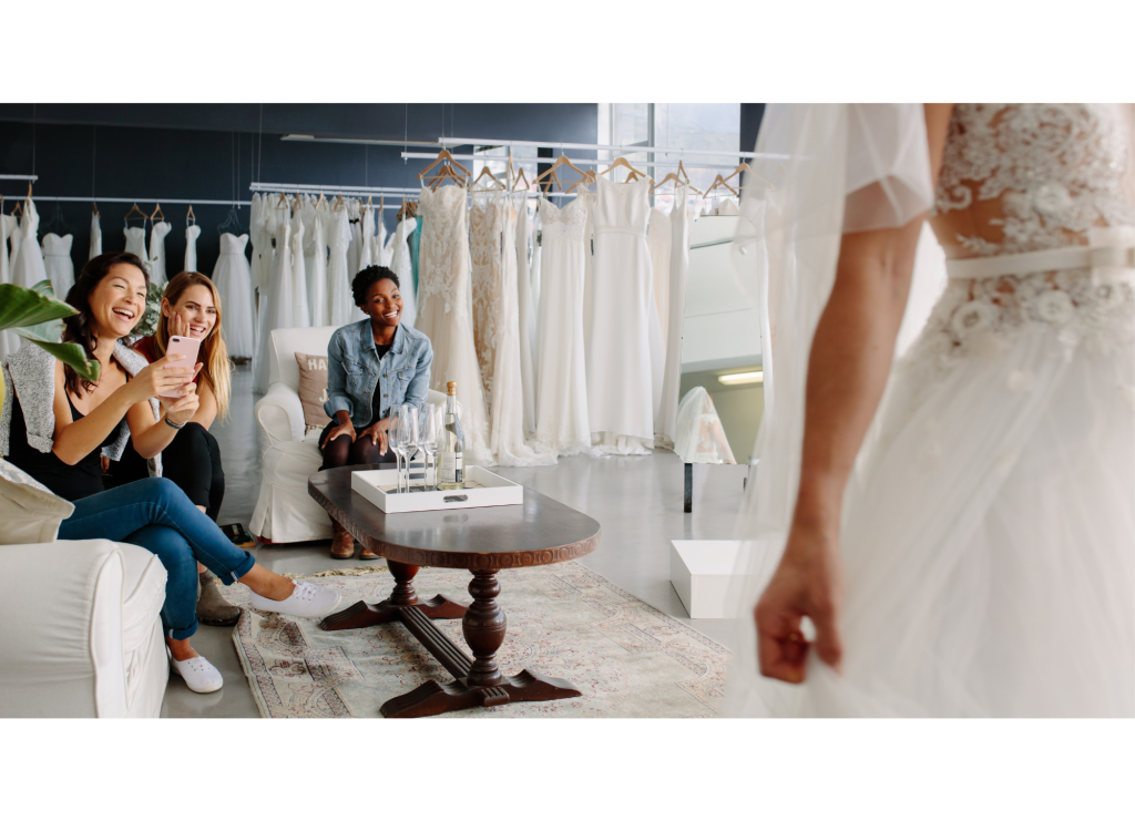 Braut probiert ein Hochzeitskleid in einem Brautmodengeschäft an, während ihre Freundinnen auf einem Sofa sitzen, lächeln und mit dem Handy Fotos machen. Im Hintergrund hängen elegante Brautkleider an Kleiderbügeln.