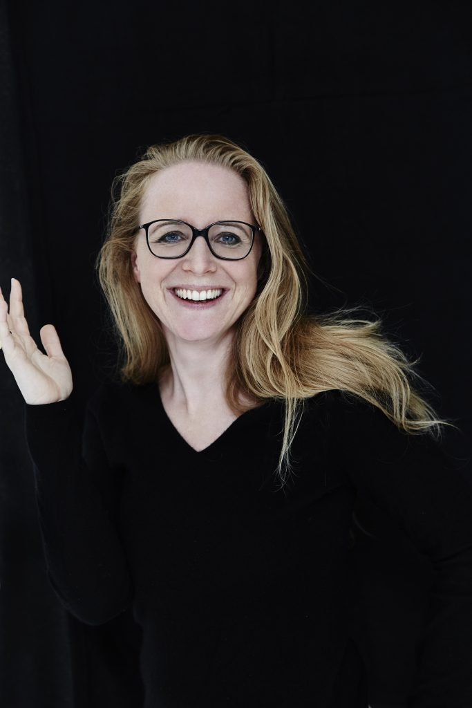 Porträt einer lächelnden Frau mit langen, welligen blonden Haaren und schwarzer Brille. Sie trägt ein schwarzes Oberteil und hebt freundlich eine Hand. Der Hintergrund ist schwarz.