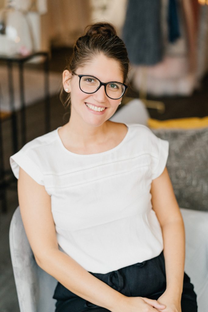 Lächelnde Frau mit dunkler Brille und hochgestecktem Haar sitzt entspannt in einem eleganten Brautmodengeschäft. Sie trägt eine weiße Bluse und schwarze Hose. Im Hintergrund sind Kleider und stilvolle Dekorationselemente zu sehen.