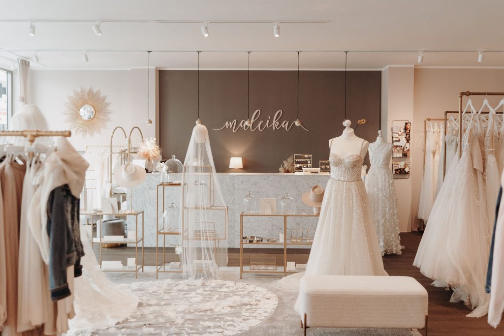 Eleganter Brautmoden-Store mit stilvollem Interieur in sanften Beige- und Goldtönen. Im Vordergrund stehen eine Schaufensterpuppe mit glitzerndem Brautkleid, ein langer Schleier auf einer Halterung sowie dekorative Elemente wie Hüte und Schmuck. Im Hintergrund ist eine stilvolle Theke mit dem Schriftzug „maleika“ zu sehen.
