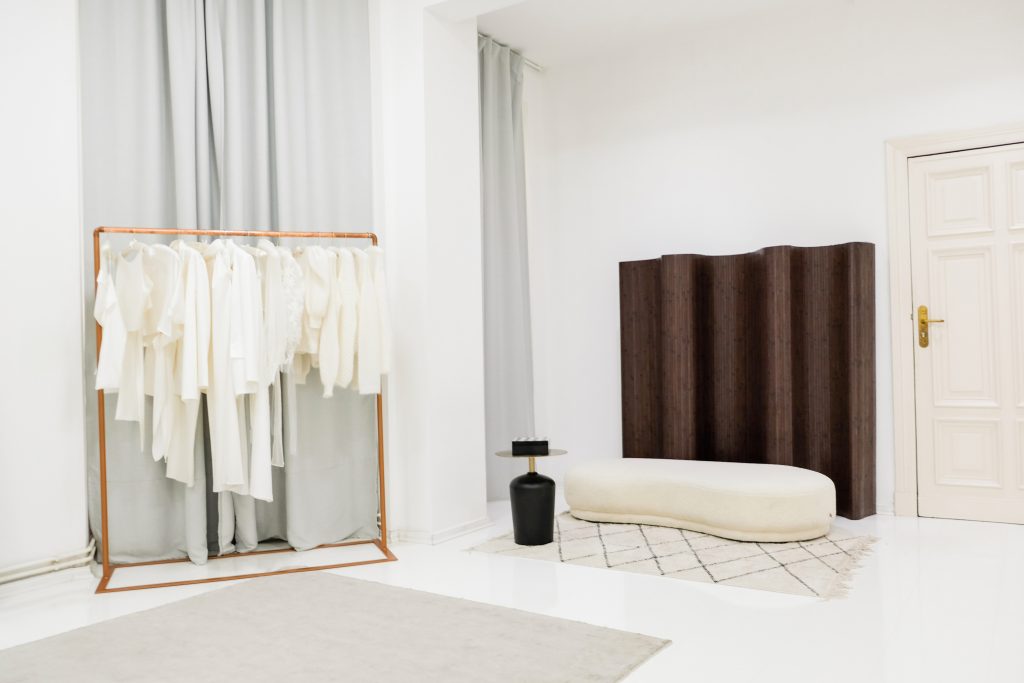 Minimalistisch eingerichteter Anproberaum in einem Brautmodengeschäft mit einer Auswahl an weißen Kleidungsstücken auf einem kupferfarbenen Kleiderständer. Ein dunkler Raumtrenner, eine elegante cremefarbene Sitzbank und dezente Teppiche schaffen eine stilvolle, ruhige Atmosphäre.