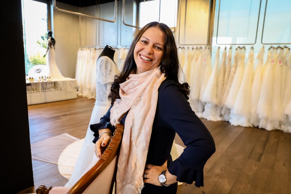 Eine lachende Frau mit langen, dunklen Haaren steht in einem eleganten Brautmodengeschäft. Sie trägt ein dunkelblaues Outfit mit einem rosafarbenen Schal und lehnt an einem stilvollen Stuhl. Im Hintergrund hängen zahlreiche weiße Brautkleider auf Kleiderstangen, während Schaufensterpuppen verschiedene Designs präsentieren.