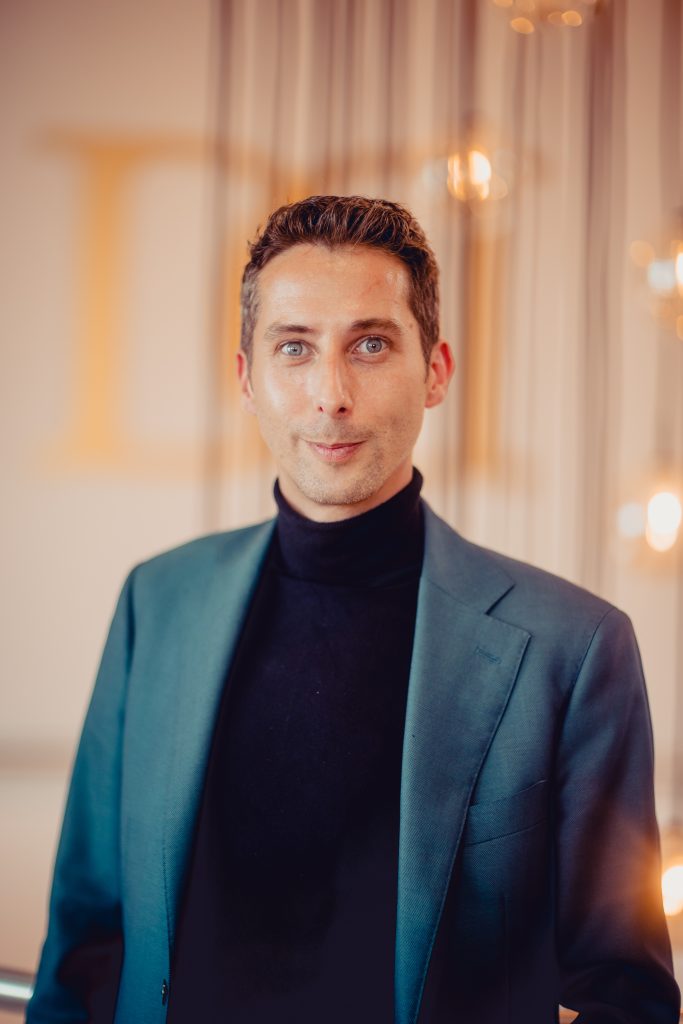 Ein Mann mit kurzen, dunklen Haaren und blauen Augen trägt einen dunkelgrünen Anzug und einen schwarzen Rollkragenpullover. Er steht vor einem eleganten, warm beleuchteten Hintergrund mit hängenden Lichtelementen und blickt direkt in die Kamera.