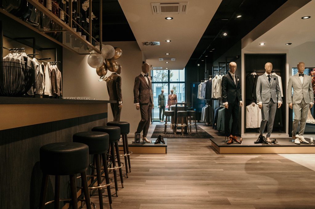 Ein stilvoll eingerichteter Herrenausstatter mit eleganten Anzügen auf Schaufensterpuppen. Im Vordergrund eine Bar mit schwarzen Hockern und goldenen Ballons, im Hintergrund eine gemütliche Lounge und große Fenster.