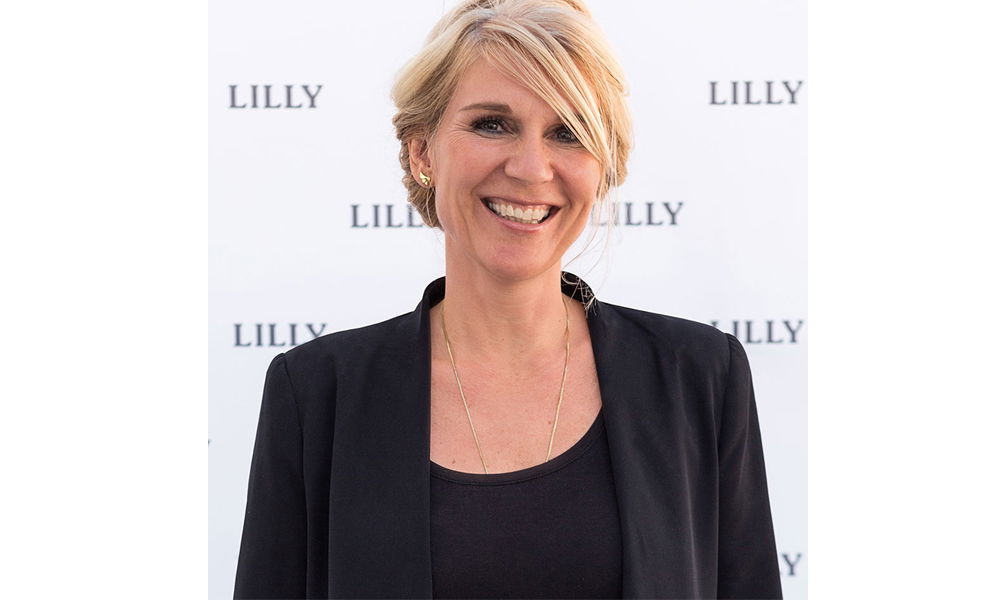 Eine lächelnde Frau mit blonden Haaren in einer eleganten schwarzen Jacke steht vor einer weißen Wand mit dem Schriftzug „LILLY“.