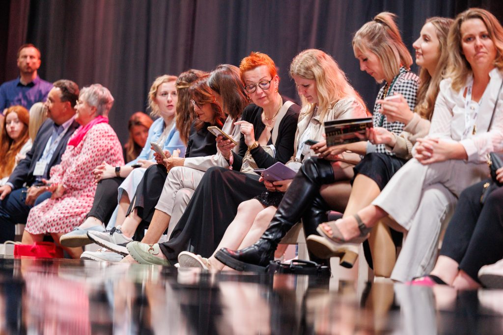 Een diverse groep vakbezoekers geniet van een modeshow op de European Bridal Week. De sfeer is gefocust, terwijl velen notities maken of foto’s nemen.