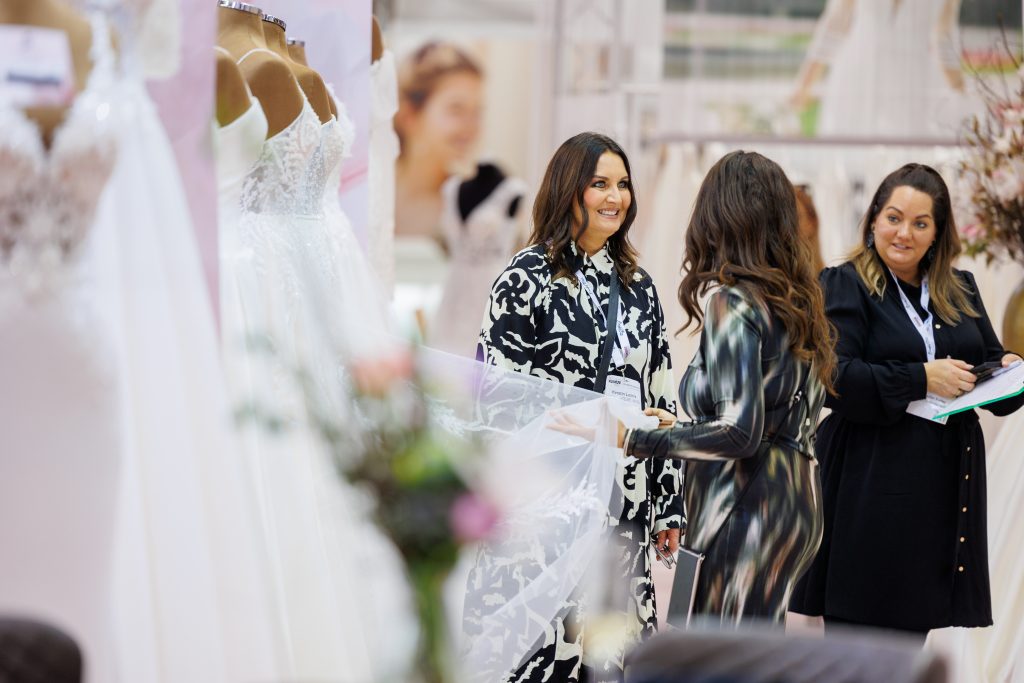 Een bruidsmode-expert bespreekt de nieuwste trouwjurkcollecties met een klant op de European Bridal Week. De prachtige jurken op de achtergrond creëren een inspirerende setting.