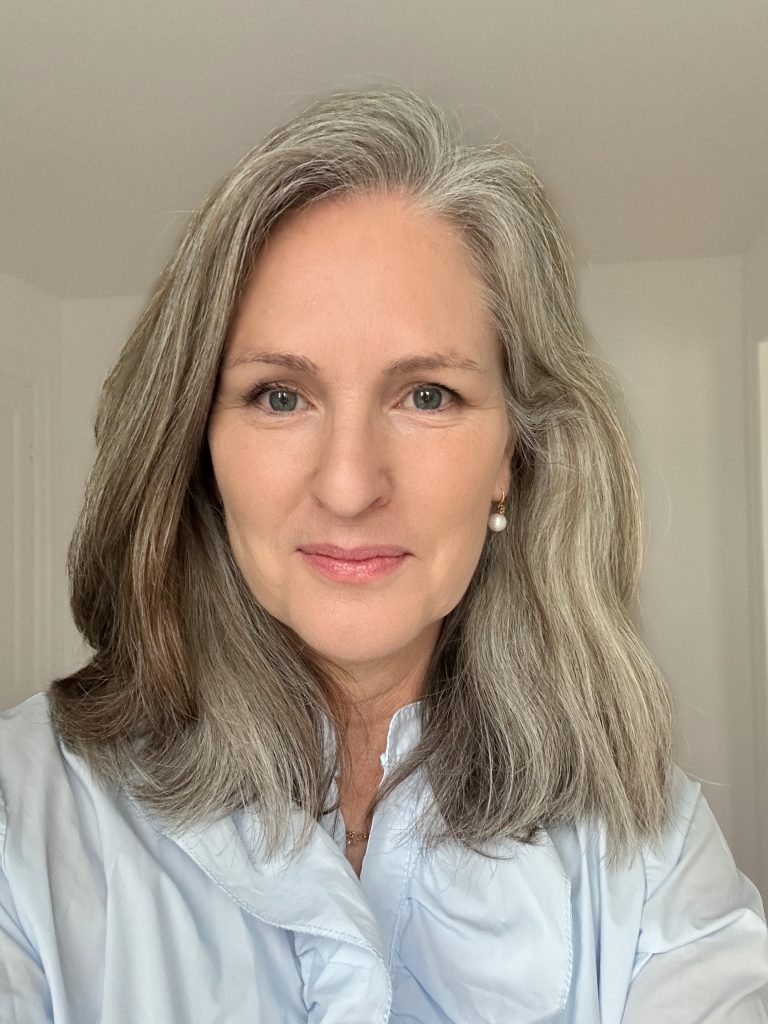Portret van een vrouw met grijs haar en een blauwe blouse die glimlachend in de camera kijkt.