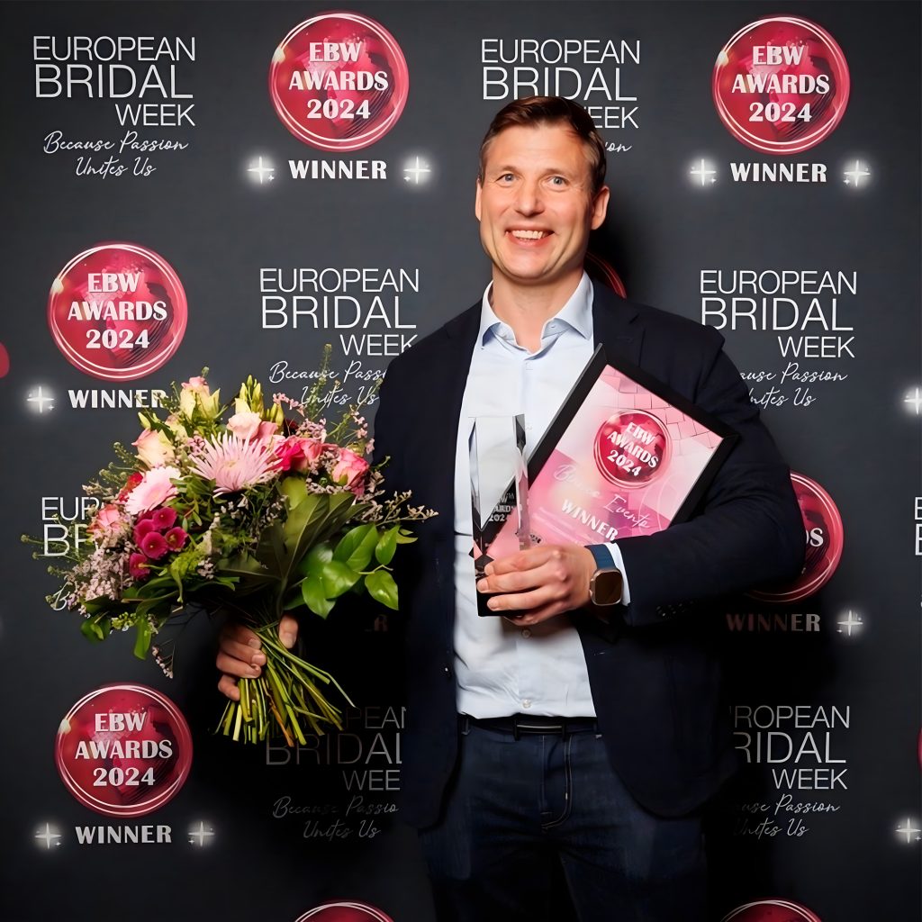Een man houdt een boeket bloemen en een winnaarscertificaat vast tijdens de European Bridal Week Awards 2024, voor een zwarte achtergrond met logo's.