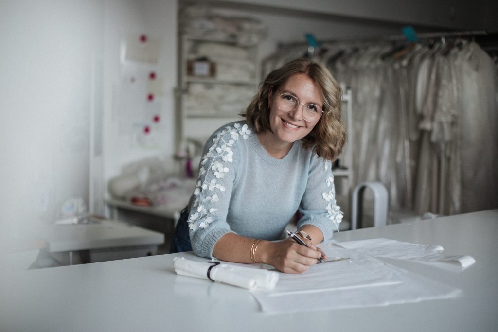 Een modeontwerpster in een lichtblauwe trui met bloemendetails zit aan een witte tafel en tekent een patroon in een atelier met bruidsjurken op de achtergrond.