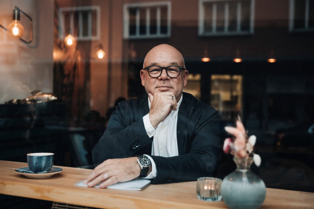 Ein nachdenklicher Mann mit Brille sitzt in einem Café und schaut durch das Fenster nach draußen. Er trägt einen dunklen Anzug mit weißem Hemd und stützt sein Kinn auf die Hand.