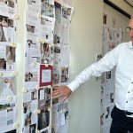 Rob Klaarenbeek steht lächelnd vor einer Wand voller Layout-Entwürfe für ein Hochzeitsmagazin und diskutiert die Seitenplanung.
