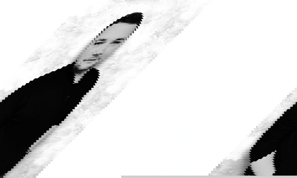 Schwarz-weißes Porträt eines Mannes mit kurzen Haaren, leicht seitlich zur Kamera stehend, vor einer marmorierten Wand.