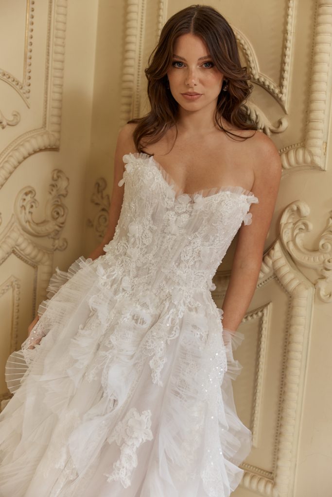 Brautmodel in einem romantischen Spitzenkleid mit 3D-Blüten und Tüll-Details vor kunstvoll verzierter Wand.