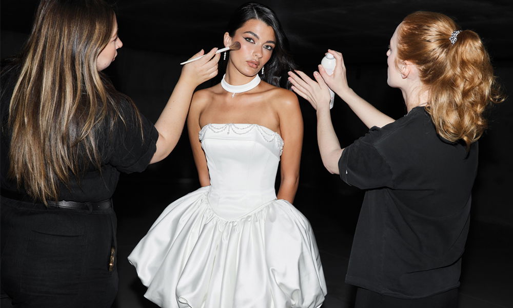 Model im modernen Mini-Brautkleid wird von zwei Stylistinnen für ein Fotoshooting geschminkt und frisiert.