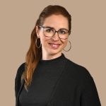 Porträt einer lächelnden Frau mit Brille und langen, rötlich-braunen Haaren im Pferdeschwanz vor beigefarbenem Hintergrund.