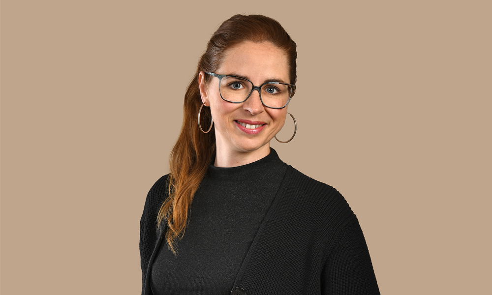 Porträt einer lächelnden Frau mit Brille und langen, rötlich-braunen Haaren im Pferdeschwanz vor beigefarbenem Hintergrund.
