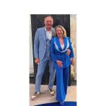 Erich und Christiane Huber stehen gemeinsam lächelnd vor einer Wand. Er trägt einen hellblauen Anzug mit Weste und Sneakers, sie ein leuchtend blaues Hosenanzug-Ensemble mit weiß-blauen Akzenten.