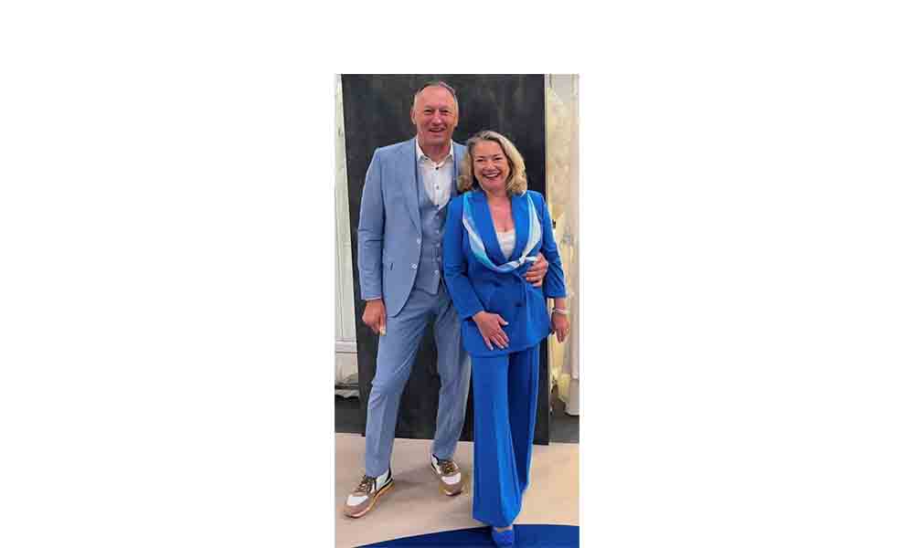 Erich und Christiane Huber stehen gemeinsam lächelnd vor einer Wand. Er trägt einen hellblauen Anzug mit Weste und Sneakers, sie ein leuchtend blaues Hosenanzug-Ensemble mit weiß-blauen Akzenten.