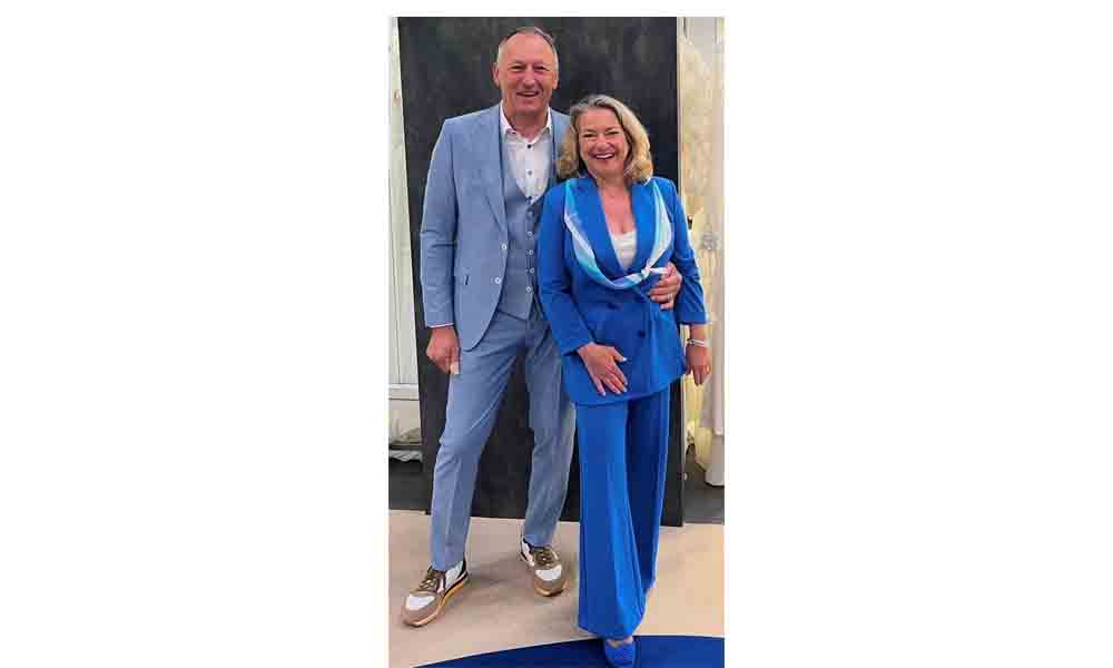 Erich und Christiane Huber stehen gemeinsam lächelnd vor einer Wand. Er trägt einen hellblauen Anzug mit Weste und Sneakers, sie ein leuchtend blaues Hosenanzug-Ensemble mit weiß-blauen Akzenten.
