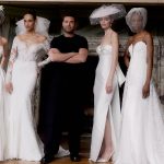Vier Models in eleganten Brautkleidern posieren neben einem Designer in schwarzem Outfit, aufgenommen vor einem Kamin mit Marmordetails.