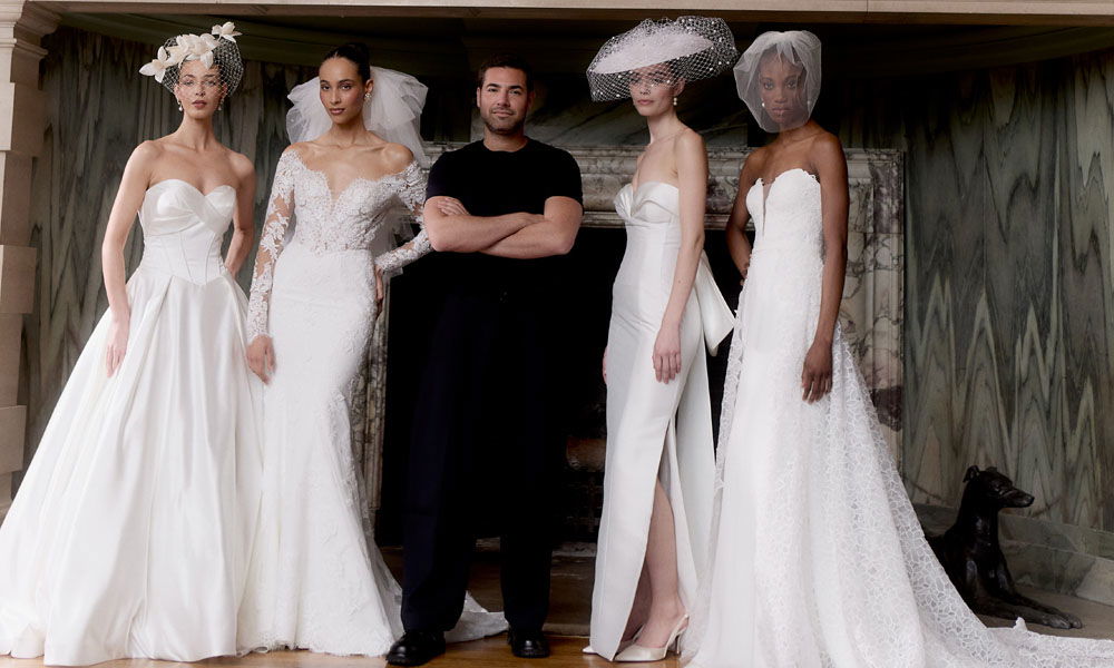 Vier Models in eleganten Brautkleidern posieren neben einem Designer in schwarzem Outfit, aufgenommen vor einem Kamin mit Marmordetails.