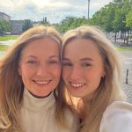 Zwei Frauen lächeln in die Kamera und machen ein Selfie im Freien, mit grünem Platz und Bäumen im Hintergrund.