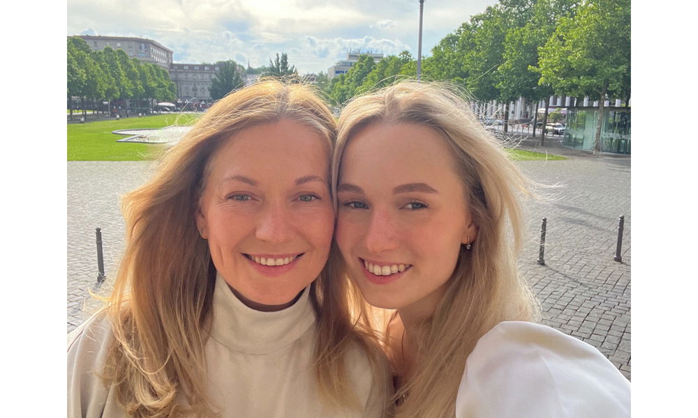 Zwei Frauen lächeln in die Kamera und machen ein Selfie im Freien, mit grünem Platz und Bäumen im Hintergrund.