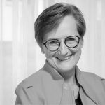 Schwarz-weiß Porträt einer lächelnden Frau mit Brille und kurzem Haar vor einem hellen Vorhang.