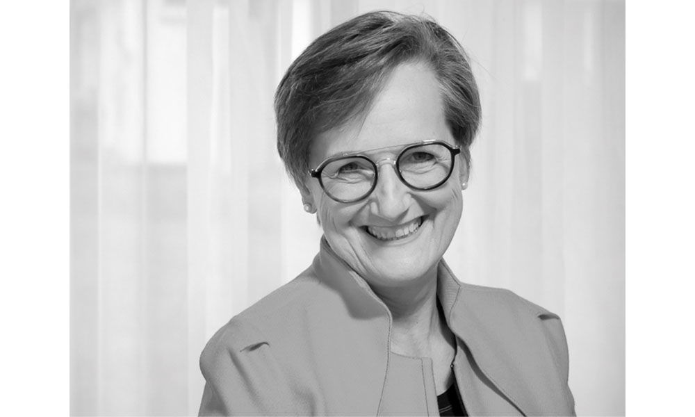 Schwarz-weiß Porträt einer lächelnden Frau mit Brille und kurzem Haar vor einem hellen Vorhang.