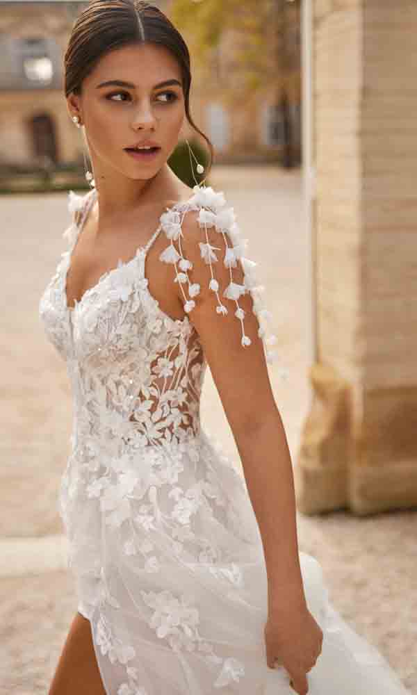 Braut in einem transparenten Spitzenkleid mit 3D-Blüten und verspielten Schulterdetails im Außenbereich.