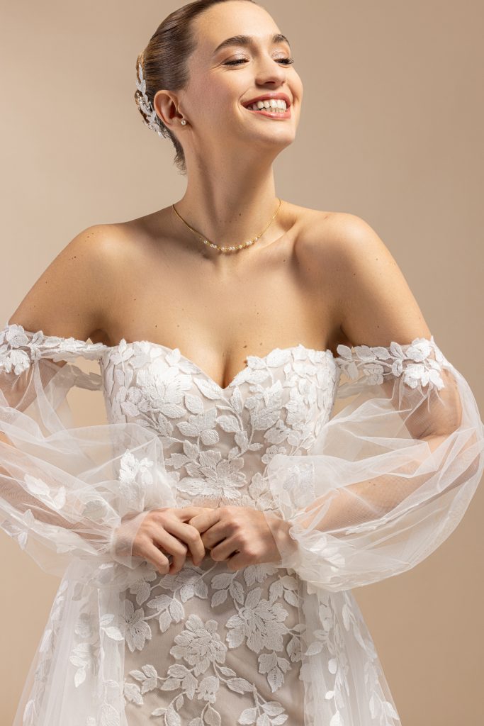 Off-shoulder bruidsjurk met bloemrijk kant en volumineuze tule mouwen, gefotografeerd tegen een rustige, neutrale achtergrond.