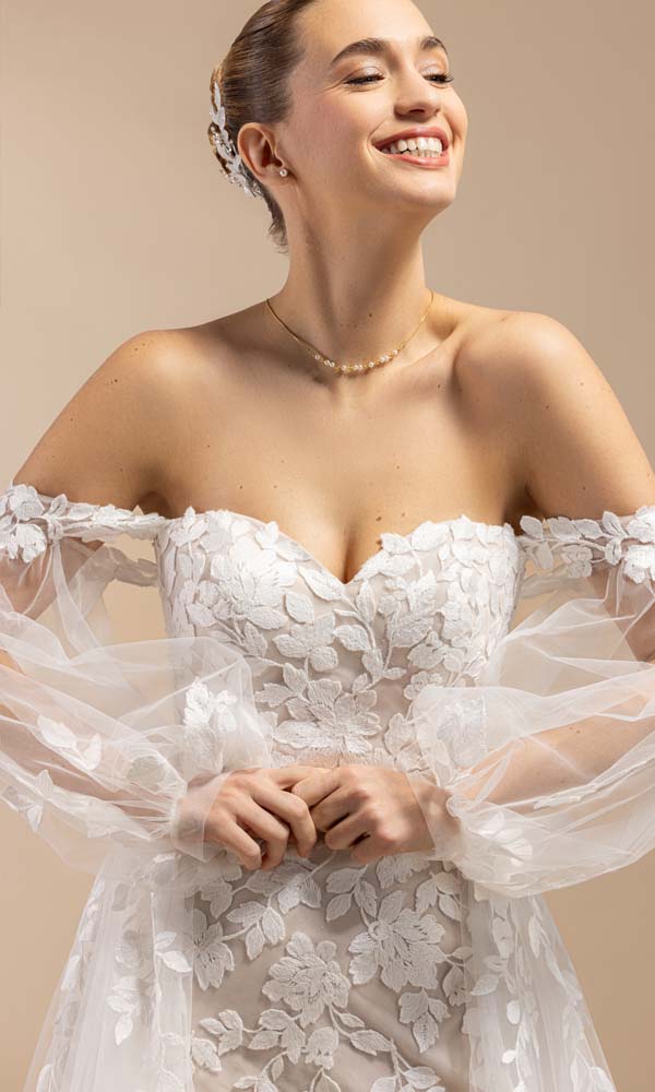 Lächelnde Braut im schulterfreien Spitzenkleid mit voluminösen Tüllärmeln und floralen Applikationen.