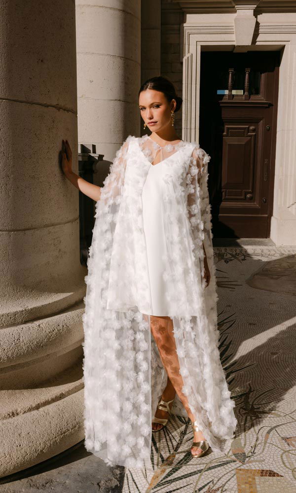 Kort bruidsjurkje gecombineerd met een opvallende cape van 3D-bloemen, gefotografeerd in een klassieke stedelijke omgeving.