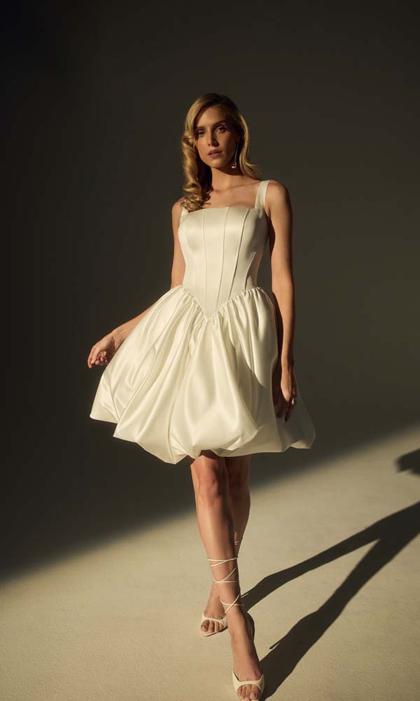 Kort satijnen bruidsjurkje met gestructureerd lijfje en speelse bubble-rok, gefotografeerd in een minimalistische studiosetting.
