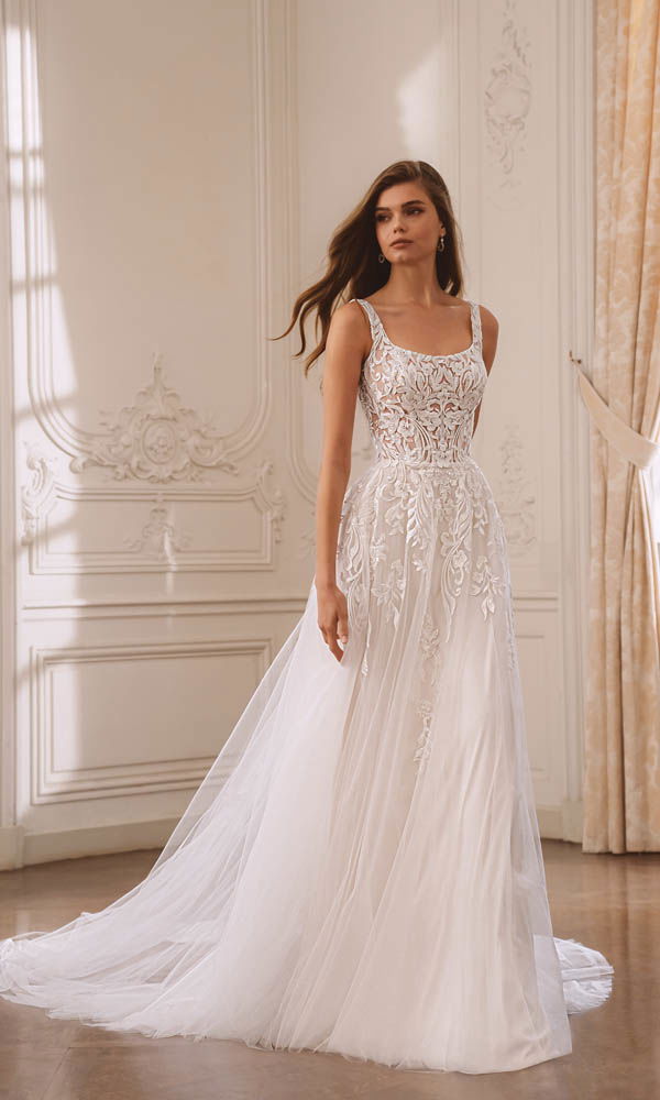 Elegante Braut in einem A-Linien-Kleid mit transparentem Spitzenoberteil und fließendem Tüllrock in einem hellen, klassischen Raum.