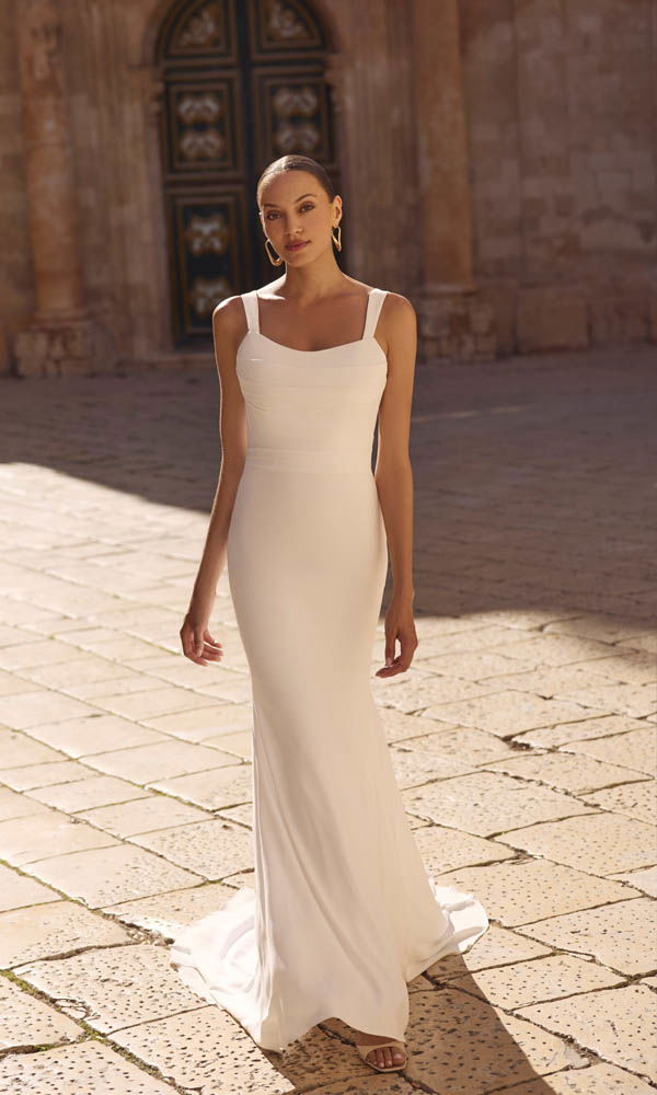 Minimalistische, slank gesneden bruidsjurk met rechte halslijn en subtiele schouderbanden, gefotografeerd in warm buitenlicht.