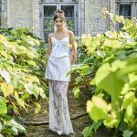 Eine Frau in einem weißen, eleganten Bridal-Look posiert in einem verwunschen wirkenden Gartenweg mit üppigem Grün.