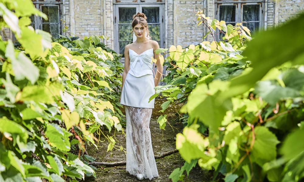 Eine Frau in einem weißen, eleganten Bridal-Look posiert in einem verwunschen wirkenden Gartenweg mit üppigem Grün.