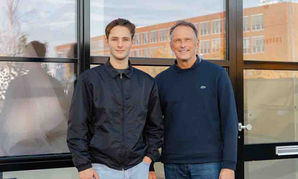 Zwei Männer stehen lächelnd vor einem modernen Gebäude mit großen Glasfronten und blicken in die Kamera.