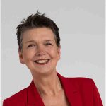 Porträt einer lächelnden Frau mit kurzem dunklem Haar in einem roten Blazer vor hellem Hintergrund.