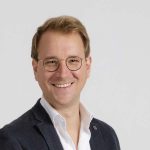 Porträt eines lächelnden Mannes mit Brille und dunklem Blazer vor neutralem Hintergrund.